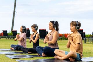 Four individuals practice yoga on mats outdoors, wearing headphones and seated in a meditative pose, surrounded by lush greenery.