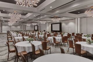 A spacious, elegant banquet hall featuring round tables with white linens, modern chandeliers, and a large presentation screen.