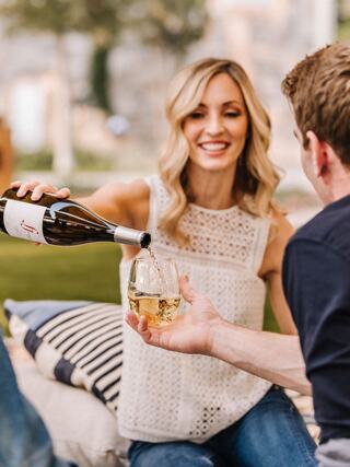 A woman joyfully pours wine into a glass for a man, both engaged in a relaxed outdoor setting. Soft sunlight and cozy cushions enhance the vibe.