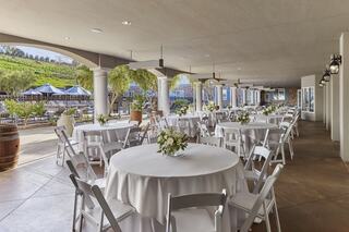 Elegant outdoor venue with circular tables, white linens, and floral centerpieces, surrounded by greenery and decorative lighting.