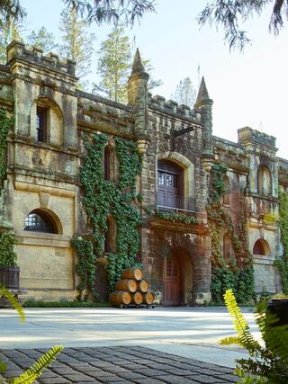 A historic stone building with ivy-covered walls and tall spires, featuring stacked barrels in front on a sunny day.
