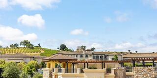 A picturesque view featuring luxury homes with stone accents, lush vineyards, and a bright blue sky dotted with clouds.