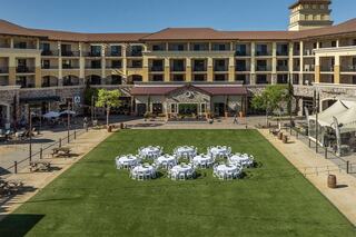A courtyard features neatly arranged tables with white cloths, surrounded by a multi-story building and lush green grass.