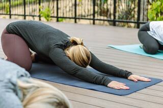 A woman practices yoga in a forward bend position on a mat, surrounded by others in a serene outdoor setting.