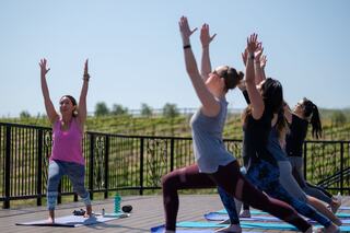A group of women practice yoga outside, with one instructor leading in a bright outfit. The setting features a scenic vineyard backdrop.