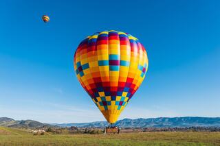 Two colorful hot air balloons float in a clear blue sky, surrounded by green hills and distant mountains, creating a vibrant scene.