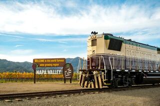 A vintage train stands on tracks beside a welcoming sign for Napa Valley, surrounded by lush vineyards under a blue sky.