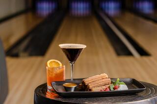 A stylish dessert platter featuring churros, sauces, and two cocktails, set against a bowling alley backdrop.