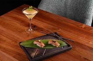A stylish presentation features two pieces of sushi on a bamboo leaf, paired with a sophisticated cocktail garnished with dill.