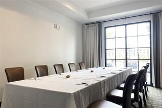 A bright meeting room with a long table covered in a white tablecloth, surrounded by brown chairs, and large windows letting in natural light.