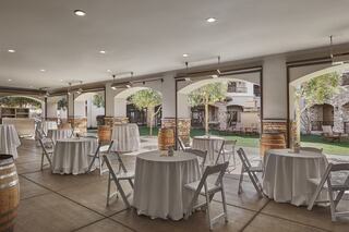 A spacious venue features round tables with white tablecloths, wooden barrels, and lush greenery visible through arched windows.