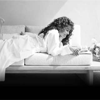 A woman lounges comfortably on a couch, absorbed in reading, dressed in a cozy robe, surrounded by soft lighting and flowers.