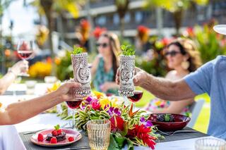 A cheerful group of friends enjoys a festive outdoor meal, raising glasses in a lush garden by a pool, surrounded by vibrant flowers.