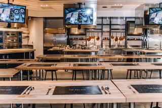 A spacious, modern kitchen equipped with multiple cooking stations, large wooden tables, and overhead screens displaying culinary content.