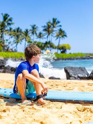 A young boy sits on a surfboard on the sandy beach, gazing at the ocean, surrounded by palm trees and clear blue skies.