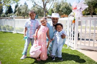 A cheerful grandfather poses with three joyful children outdoors, dressed in colorful attire, surrounded by greenery and a festive atmosphere.