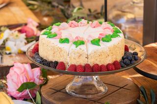 A beautifully decorated cake topped with pink and green icing, surrounded by fresh berries, sits on a glass cake stand amidst floral decor.