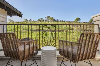 A cozy balcony features two woven chairs and a small table, overlooking lush green fields and vibrant trees under a clear blue sky.