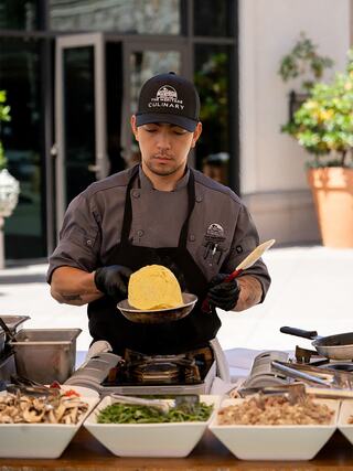 A chef in a gray uniform carefully plates a yellow dish while surrounded by a variety of fresh ingredients in an outdoor setting.