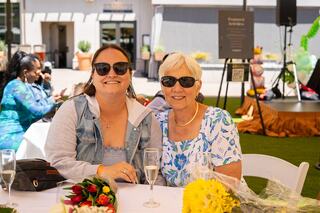 Two women sit at a table, smiling and enjoying a sunny day. Flowers are nearby, and they're wearing sunglasses in a festive setting.