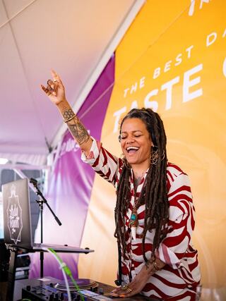 A joyful DJ with long dreadlocks, dressed in a bold red and white outfit, passionately performs at a vibrant outdoor event.