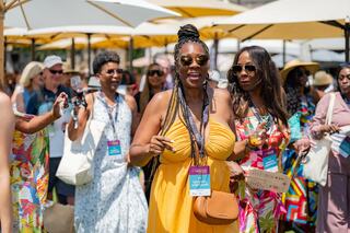 A joyful group of women, dressed in vibrant summer dresses, enjoy a sunny outdoor event, laughing and sipping drinks under umbrellas.