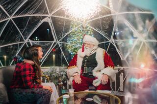 A child chats with Santa inside a cozy igloo-like space, surrounded by festive decorations and holiday cheer.