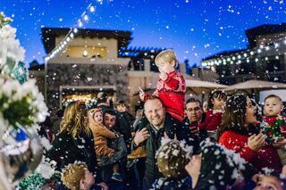 A festive gathering with people celebrating, children in holiday attire, and artificial snow falling amidst cheerful decorations and lights.