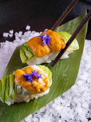 Delicate sushi topped with bright orange uni and avocado, garnished with purple flowers, presented on a bed of coarse salt.
