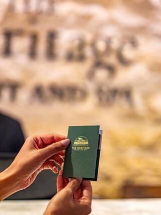 A person holds a green booklet with a logo, sitting at a reception desk with a blurred background featuring the name of a resort and spa.