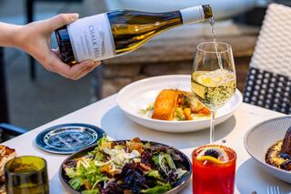 A hand pours white wine into a glass, surrounded by a vibrant salad, gourmet dish, and a colorful cocktail on a dining table.