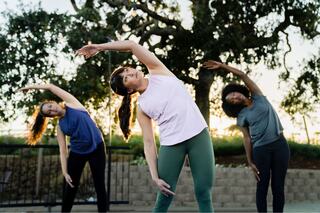 Three women practice side stretches in a park during sunset, showcasing fitness and movement amidst a scenic backdrop of trees.