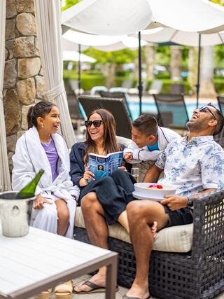 A family enjoys a joyful moment by the pool, laughing and sharing snacks while relaxing in cozy seating on a sunny day.