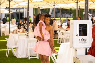 A woman in a pink dress holds a young girl in a matching dress, smiling at an outdoor event filled with white tables and umbrellas.