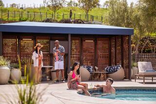 Four friends enjoy a sunny day poolside, sipping drinks and relaxing in a stylish outdoor setting with lush greenery in the background.