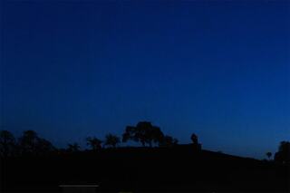 A serene duskscape featuring silhouettes of trees and a figure atop a hill against a deep blue sky, evoking tranquility and mystery.