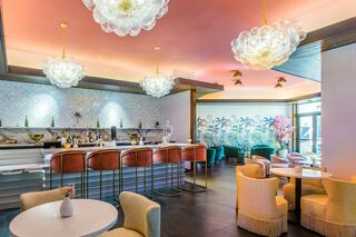 A stylish bar area features elegant seating, modern decor, and vibrant plants, illuminated by unique light fixtures and soft pastel tones.