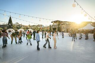 A lively outdoor ice skating rink features people enjoying the activity, with a festive tree and lights under a warm sunset glow.