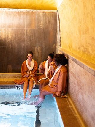 Three women in cozy bathrobes relax by a tranquil pool, enjoying drinks and laughter in a serene wellness setting.