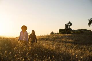 Two girls walk through tall grass at sunset, with a distant statue silhouetted against the warm orange sky.
