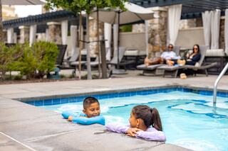Two children chat playfully in a pool, while adults relax nearby. Lush greenery and lounge chairs create a serene atmosphere.