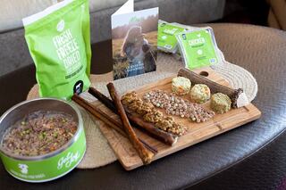 A wooden platter displays various pet treats, including freeze-dried food, flavored bites, and chew sticks, alongside a promotional booklet.