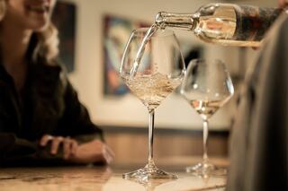A person pours white wine into a tall glass, while another individual enjoys the moment at a bar, surrounded by artistic decor.
