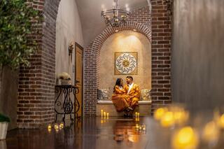 A couple in matching outfits sit closely in a cozy, warmly lit hallway adorned with candles and brick walls, creating an intimate atmosphere.