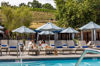 A couple enjoys drinks by a poolside with a relaxed atmosphere, surrounded by umbrellas and a scenic vineyard in the background.