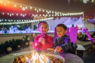 Two children roast marshmallows over a fire pit, surrounded by festive lights and holiday decor, creating a cozy and joyful atmosphere.