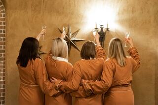 Four women in cozy robes raise champagne glasses in celebration, facing a textured wall adorned with star decorations and warm lighting.