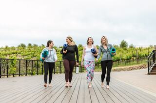 Four women walk barefoot on a wooden deck, holding yoga mats, laughing, with a vineyard backdrop and an overcast sky.