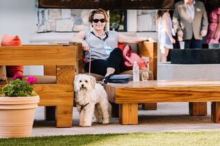 A woman relaxes on a wooden bench with a small dog beside her, enjoying a sunny day in a vibrant outdoor setting.